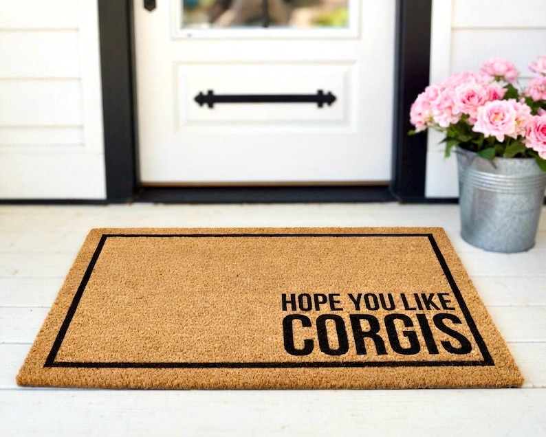 May include: A rectangular doormat made of natural coir fibre, featuring a black border and the text "HOPE YOU LIKE CORGIS" in bold, black letters. The mat is placed in front of a white door.