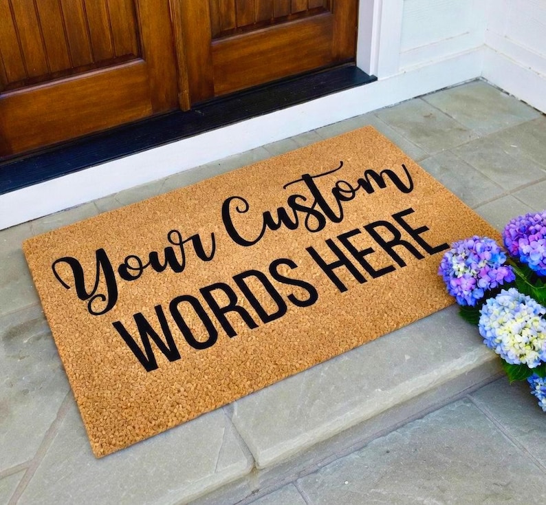 May include: A rectangular doormat made of natural coir fiber, featuring the text "Your Custom WORDS HERE" in black script. The mat is placed on a stone step near a wooden door, with colorful flowers to the right.