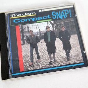 May include: A CD case featuring the album "Snap!" by The Jam. The cover art displays the band members walking on a cobblestone street. The album title and band name are in bold, colorful text.