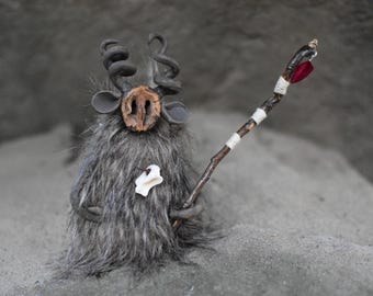 May include: A whimsical, handmade figurine of a creature with gray fur, dark curly horns, and a wooden nose. It holds a staff adorned with white bands and a red feather. The figure is set against a textured gray backdrop.