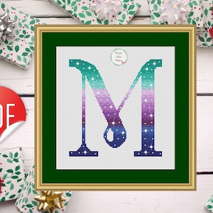 Könnte beinhalten: Kreuzstich-Design des Buchstabens "M" in einem goldenen Rahmen. Der Buchstabe weist einen Farbverlauf aus Türkis, Lila und Blau mit weißen Punktakzenten auf. Der Hintergrund ist weiß und der Rahmen grün. Das Bild ist von verpackten Geschenken umgeben.