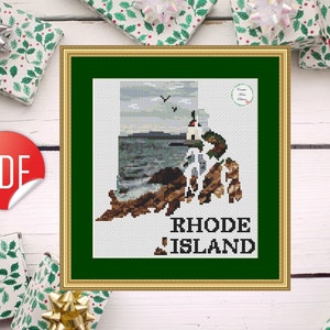 May include: Framed cross-stitch art featuring the outline of Rhode Island, with a lighthouse and coastal scene. The text "RHODE ISLAND" is stitched below the image. The artwork is in a gold frame with a green mat, surrounded by wrapped gifts.