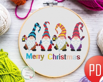 May include: A cross-stitch pattern in a wooden embroidery hoop featuring four gnomes with colorful hats and beards, with the text Merry Christmas in multi-colored letters below them. The design is displayed on a white fabric background.