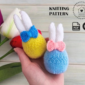 Puede incluir: Dos adornos de conejito tejidos, uno amarillo y otro azul, cada uno con orejas blancas y una pajarita. El texto "KNITTING PATTERN" se muestra encima de un logotipo. Hay tulipanes en el fondo.