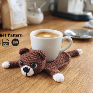 May include: A brown crocheted bear coaster with white accents, holding a white coffee cup. The coaster is handmade and designed to protect surfaces from heat and spills. The image also includes the text "Crochet Pattern".