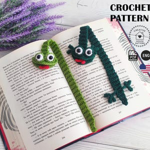 May include: Crocheted frog bookmarks in green with white eyes and red tongues rest on an open book. The text "CROCHET PATTERN" is visible. The bookmarks are handmade and add a whimsical touch to reading.