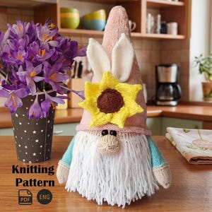 Puede incluir: Una muñeca gnomo tejida con un gorro rosa, barba blanca y mangas azules. El gnomo tiene orejas de conejo y un girasol amarillo. Un jarrón de flores moradas está a la izquierda. El texto "Knitting Pattern" es visible.
