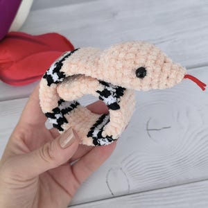 May include: A pink crochet pattern snake slap bracelet with black and white checkered accents. The snake has a red tongue and a black eye.