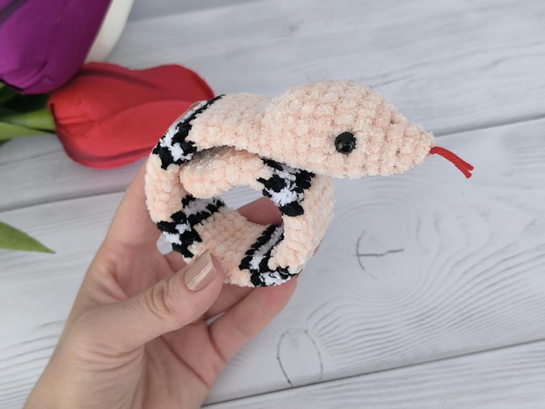 May include: A pink crochet pattern snake slap bracelet with black and white checkered accents. The snake has a red tongue and a black eye.