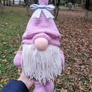 May include: A handmade crochet gnome doll with a pink hat and feet, a beige beard, and a round nose. The gnome has a silver bow on its hat. The doll is held in front of a blurred background of trees and fallen leaves.