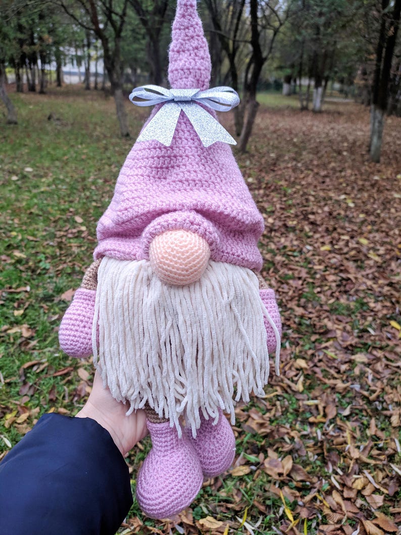 May include: A handmade crochet gnome doll with a pink hat and feet, a beige beard, and a round nose. The gnome has a silver bow on its hat. The doll is held in front of a blurred background of trees and fallen leaves.