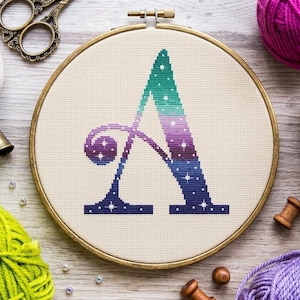 Könnte beinhalten: Ein Kreuzstich-Stickrahmen mit dem Buchstaben "A" in einem Farbverlauf aus Türkis, Lila und Marineblau mit weißen Sternakzenten. Die Stickerei befindet sich in einem Holzrahmen, umgeben von Bastelmaterialien wie Garn und Schere.