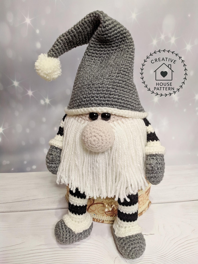 A crochet pattern gnome doll with a gray hat and body, white beard, and pink nose. The gnome has black and white striped legs and is wearing gray shoes.