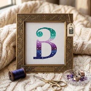 Puede incluir: Punto de cruz de la letra "B" en un marco dorado decorativo. La letra presenta un degradado de colores que va del verde azulado al morado y al azul oscuro, con acentos de estrellas blancas. Una etiqueta dice "PDF ENG". También hay un carrete de hilo morado, tijeras y nociones de costura.
