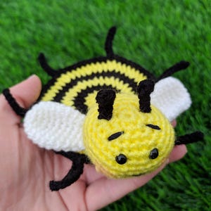 May include: A handmade crochet bee mug rug. The bee features a yellow head and body with black stripes, black antennae, and black eyes. The wings are white. The toy is held in a hand against a green background.