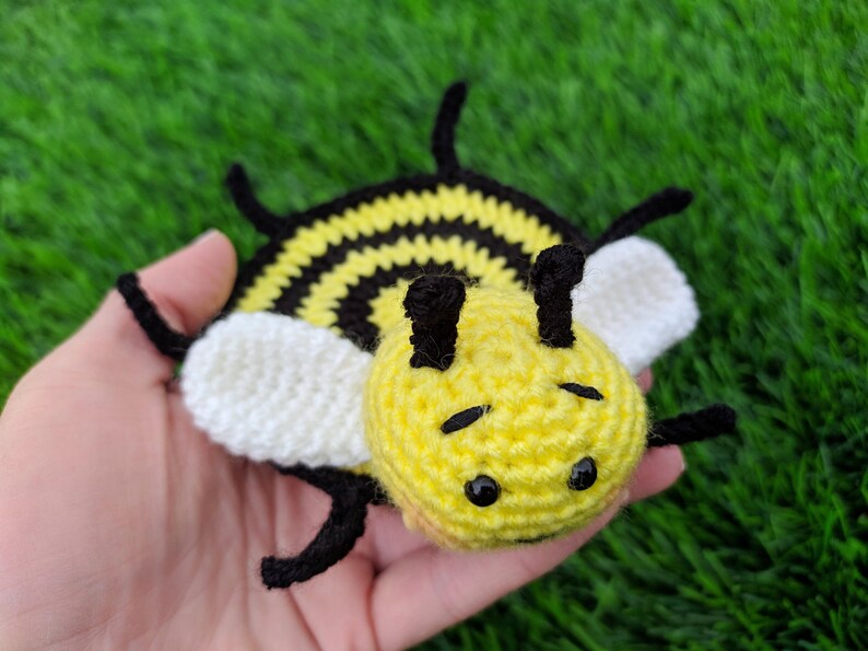 May include: A handmade crochet bee mug rug. The bee features a yellow head and body with black stripes, black antennae, and black eyes. The wings are white. The toy is held in a hand against a green background.