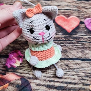 Peut inclure: Poup&eacute;e chat grise au crochet avec un n&oelig;ud orange, des yeux noirs et un nez rose. Le chat porte une robe verte et orange. La poup&eacute;e est tenue par une main. Un c&oelig;ur rose et le mot "love" sont en arri&egrave;re-plan.