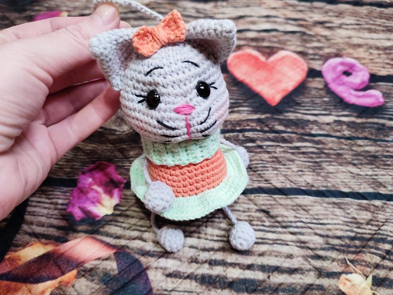 Peut inclure: Poup&eacute;e chat grise au crochet avec un n&oelig;ud orange, des yeux noirs et un nez rose. Le chat porte une robe verte et orange. La poup&eacute;e est tenue par une main. Un c&oelig;ur rose et le mot "love" sont en arri&egrave;re-plan.