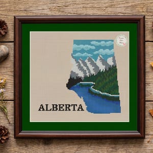May include: A framed cross-stitch embroidery featuring a landscape of Alberta, Canada. The design shows snow-capped mountains, a dense green forest, and a blue lake within the province's shape on a beige background, with the text ALBERTA below.