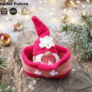 May include: A handmade crochet gnome in a pink basket. The gnome has a red hat with a white flower and a white beard. The basket is pink and tan with white butterfly accents. The image includes the text "Crochet Pattern".
