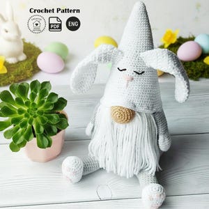 May include: A crocheted Easter gnome with bunny ears, a long white beard, and closed eyes. The gnome is in shades of grey and white, with a tan nose and a white beard. The background includes a succulent plant, Easter eggs, and a ceramic bunny.
