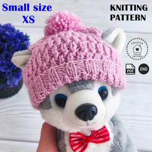 Può includere: Un cappello rosa lavorato a maglia con un pompon, etichettato "Small size XS", è posto su un giocattolo di peluche a forma di husky. L'immagine include anche il testo "KNITTING PATTERN" e un logo con icone "PDF" e "ENG", che indica un modello per il cappello.