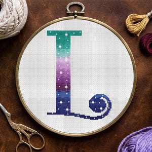 May include: A cross-stitch embroidery hoop featuring the letter "L" in a gradient of teal, purple, and blue, with white star accents. The design is set against a white fabric, framed by a brass hoop. Craft supplies like yarn and scissors are visible.