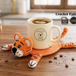 May include: A cream-colored mug with coffee rests on an orange crochet tiger coaster. The coaster has black stripes and white accents. The mug has a black logo that says "Creative House Pattern". The image also includes the text "Crochet Pattern".