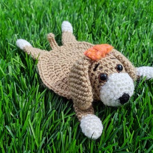 A handmade crochet dog coaster in a lying-down pose. The coaster is primarily tan with white paws and a white face. It has a small orange bow and black eyes.