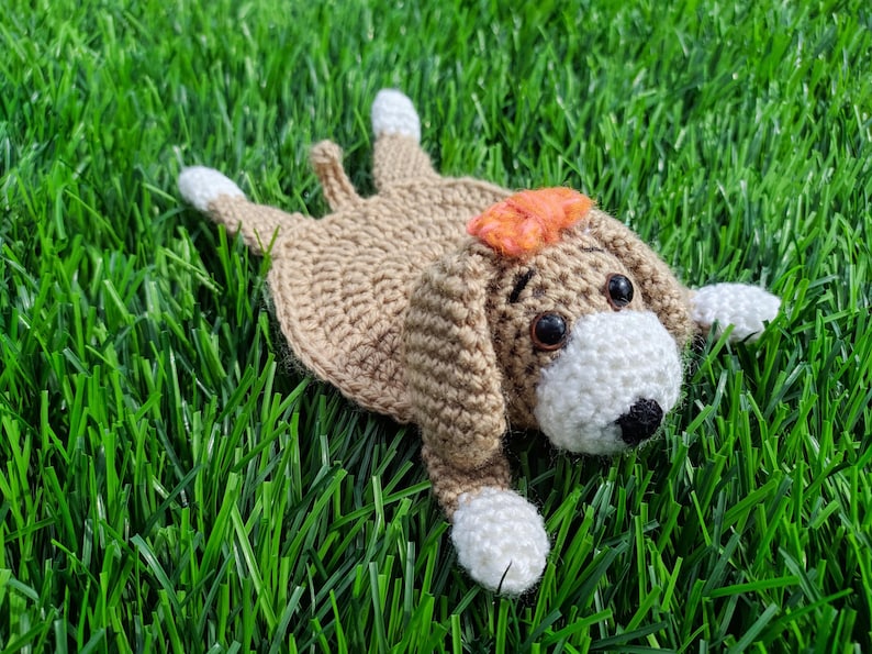 A handmade crochet dog coaster in a lying-down pose. The coaster is primarily tan with white paws and a white face. It has a small orange bow and black eyes.