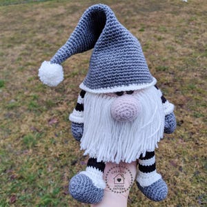 May include: A handmade crochet gnome doll with a long gray hat, white beard, and a pink nose. The gnome has striped arms and legs, and a white pom-pom on the hat. The doll is held in a hand, and the background is blurred.