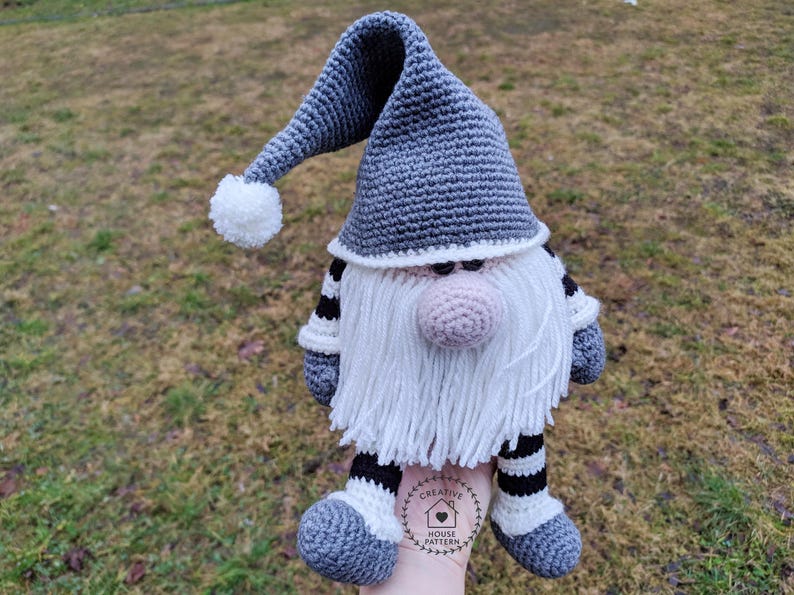 May include: A handmade crochet gnome doll with a long gray hat, white beard, and a pink nose. The gnome has striped arms and legs, and a white pom-pom on the hat. The doll is held in a hand, and the background is blurred.