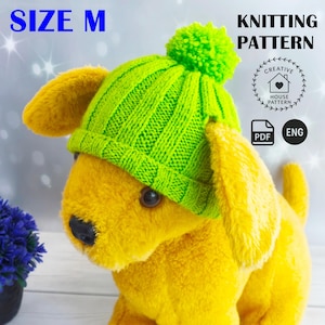 Op de afbeelding: Een gele pluchen hond draagt een felgroene gebreide muts met een pompon. De muts is gemarkeerd met "SIZE M" en de afbeelding bevat de tekst "KNITTING PATTERN" met een logo en "PDF ENG".