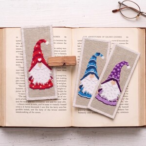 May include: Handmade gnome bookmarks with cross-stitch designs. One bookmark features a red gnome with a white beard and hat. Two others have blue and purple gnomes. The bookmarks are on a book.