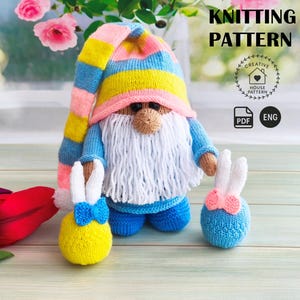 Pode incluir: Um gnomo de malha com uma longa barba branca, usando um suéter azul e um chapéu listrado em rosa, amarelo e azul. Dois enfeites de coelho de malha com laços estão em ambos os lados do gnomo. A imagem inclui o texto "KNITTING PATTERN".
