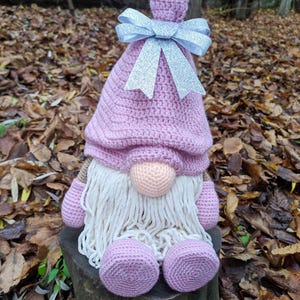 May include: A handmade crochet gnome doll with a pink hat and body, a peach-colored nose, and a long white beard. The gnome has a silver glitter bow on its hat and is sitting on a wooden stump. The background is out of focus.