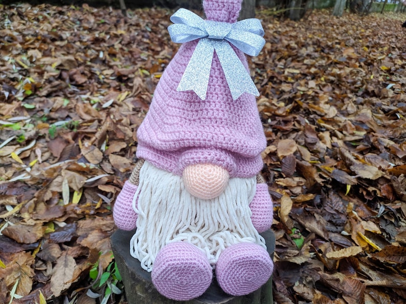May include: A handmade crochet gnome doll with a pink hat and body, a peach-colored nose, and a long white beard. The gnome has a silver glitter bow on its hat and is sitting on a wooden stump. The background is out of focus.