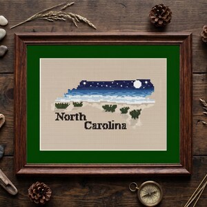 May include: A framed cross-stitch embroidery featuring the state of North Carolina in a night sky design. The map is filled with a dark blue starry sky, a white moon, and green landscape elements on a beige background with the text North Carolina.