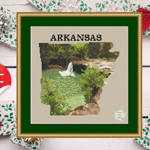May include: A cross-stitch pattern of the state of Arkansas, featuring a scenic landscape with a waterfall and greenery. The pattern is framed in a gold frame with a green mat. The title "Arkansas" is printed above the pattern.