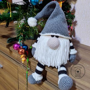 May include: A handmade crochet gnome doll with a gray pointed hat, white beard, and a beige nose. The gnome has gray and white striped legs and arms. A white pom-pom adorns the hat. The gnome is a Christmas decoration.