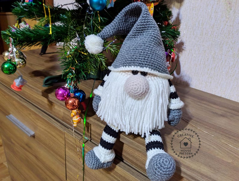 May include: A handmade crochet gnome doll with a gray pointed hat, white beard, and a beige nose. The gnome has gray and white striped legs and arms. A white pom-pom adorns the hat. The gnome is a Christmas decoration.