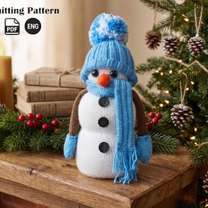 Puede incluir: Un muñeco de nieve tejido con gorro, bufanda y mitones azules, y un pompón. El muñeco de nieve tiene una nariz de zanahoria, ojos de botón negros y botones en la parte delantera. El texto "Knitting Pattern" es visible.