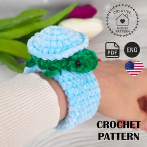 以下が含まれることがあります： 水色と緑色のカギ針編みのカメのリストバンド。カメは水色の甲羅と緑色の体を持っています。リストバンドは柔らかく、ふわふわした素材です。「CROCHET PATTERN」という文字も含まれています。