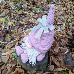 May include: A handmade pink crochet gnome doll with a long white beard, pink nose, and matching pink shoes. The gnome wears a pink hat with a silver glitter bow. The doll is sitting on a tree stump surrounded by fallen leaves.