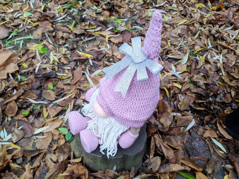 May include: A handmade pink crochet gnome doll with a long white beard, pink nose, and matching pink shoes. The gnome wears a pink hat with a silver glitter bow. The doll is sitting on a tree stump surrounded by fallen leaves.