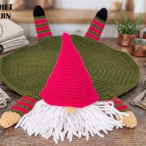 Puede incluir: Un gnomo de ganchillo con un gorro rojo brillante, barba blanca y cuerpo verde. El gnomo tiene brazos y piernas a rayas. La imagen también muestra un salvamanteles verde y el texto "CROCHET PATTERN".
