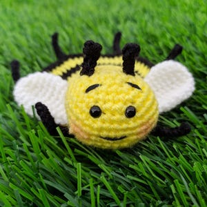 May include: A handmade crochet bee coaster with a yellow body, black stripes, and white wings. The bee has black eyes and antennae. The toy is lying on a bed of green artificial grass.