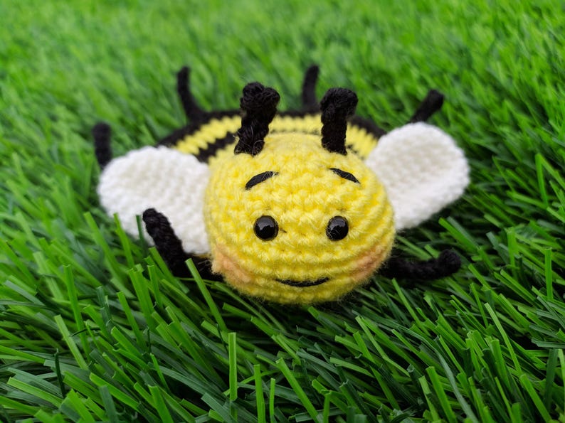 May include: A handmade crochet bee coaster with a yellow body, black stripes, and white wings. The bee has black eyes and antennae. The toy is lying on a bed of green artificial grass.
