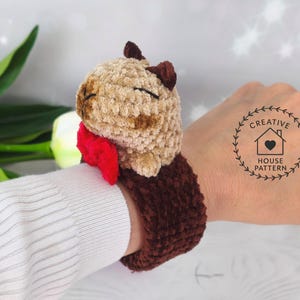 Sensory Capybara Fidget Toy Crochet Pattern wristband with a red bow tie. The Capybara has closed eyes, and the wristband is a dark brown color. The wristband is designed to be worn on the wrist.