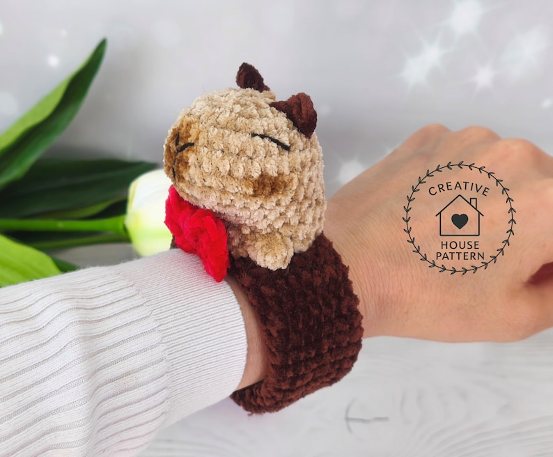 Sensory Capybara Fidget Toy Crochet Pattern wristband with a red bow tie. The Capybara has closed eyes, and the wristband is a dark brown color. The wristband is designed to be worn on the wrist.